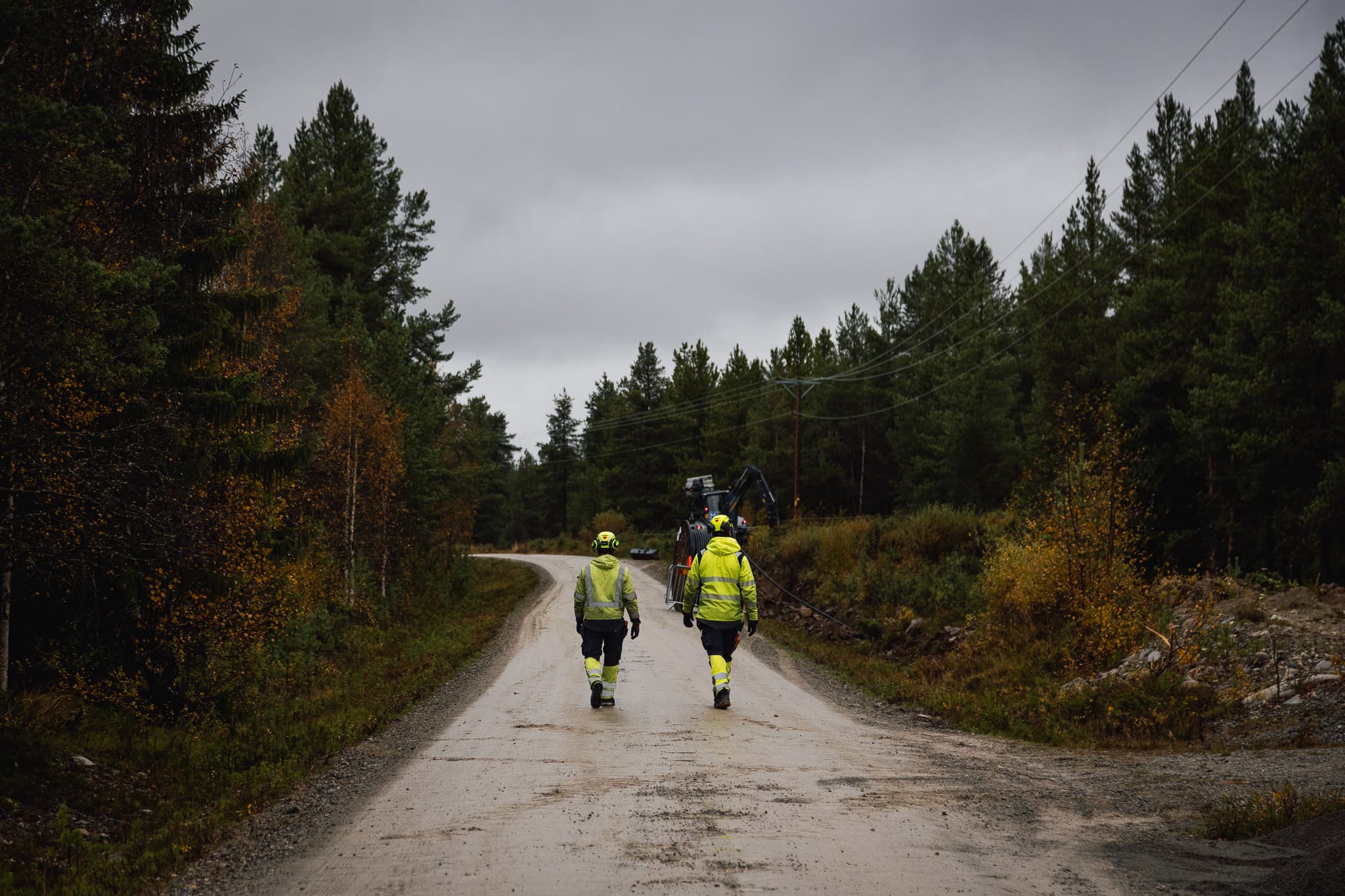 Personal från BTEA i varselkläder går längs en väg mot en traktorgrävare under elledningar, som en del av elnätsunderhåll i Jämtland.